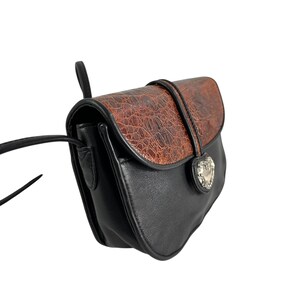 Op de afbeelding: Zwarte en bruine crossbodytas met een textuur, faux-krokodillenklep. De tas heeft een zilverkleurige hartvormige sluiting en een zwarte riem. De tas is gemaakt van leerachtig materiaal.