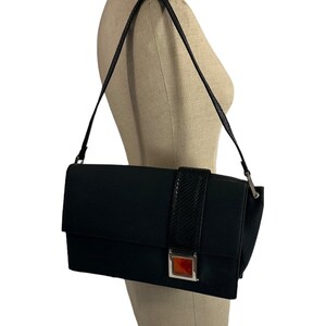 May include: A black shoulder bag with a rectangular shape. The bag features a textured black strap and a decorative band with a snakeskin pattern. A silver-toned clasp with an orange accent adds a pop of color. The bag is displayed on a mannequin.