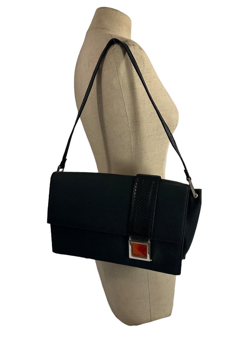 May include: A black shoulder bag with a rectangular shape. The bag features a textured black strap and a decorative band with a snakeskin pattern. A silver-toned clasp with an orange accent adds a pop of color. The bag is displayed on a mannequin.
