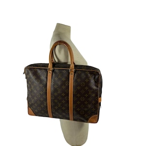 Może przedstawiać: Brązowa skórzana teczka z monogramem i górnym uchwytem. Teczka ma zamek błyskawiczny i przednią kieszeń.