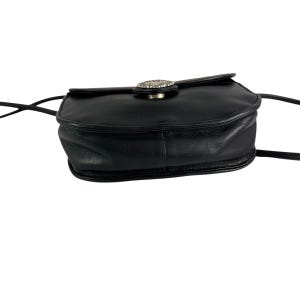 Op de afbeelding: Zwarte leren crossbodytas met een zilverkleurige decoratieve sluiting. De tas heeft een afgeronde vorm en een lange, dunne riem. De sluiting is cirkelvormig met een decoratief ontwerp. De tas is gemaakt van glad, glanzend leer.