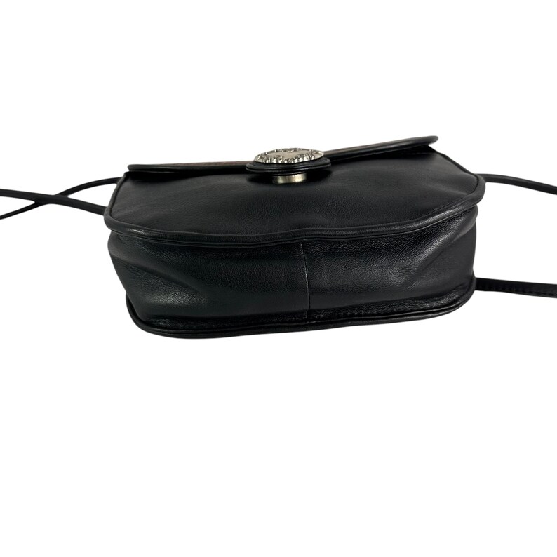 Op de afbeelding: Zwarte leren crossbodytas met een zilverkleurige decoratieve sluiting. De tas heeft een afgeronde vorm en een lange, dunne riem. De sluiting is cirkelvormig met een decoratief ontwerp. De tas is gemaakt van glad, glanzend leer.