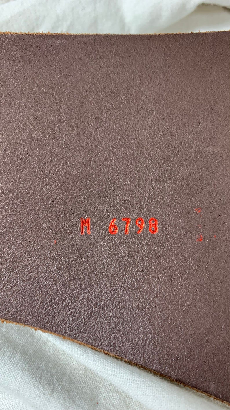 May include: Close-up of a brown leather belt with the text "M 6798" embossed in red.