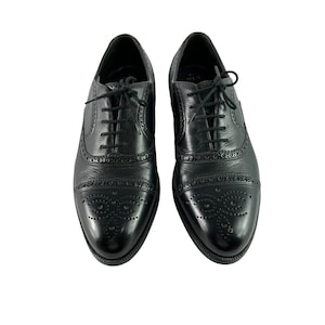Może przedstawiać: Para czarnych skórzanych butów wizytowych o klasycznym kroju Oxford. Buty posiadają noski, ozdobne perforacje i czarne sznurowadła. Buty są prezentowane na białym tle.