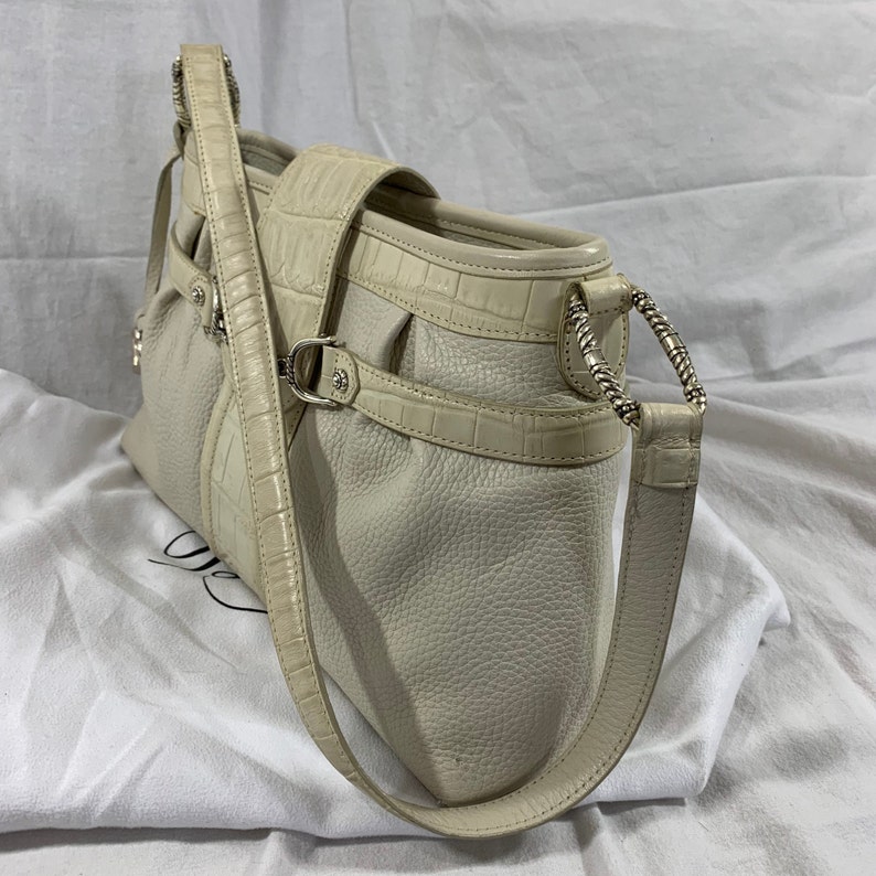 May include: A white leather handbag with a crocodile embossed design. The bag has a long strap and a silver buckle.