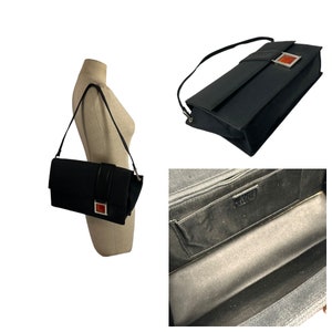 May include: Black shoulder bag with a rectangular shape. The bag features a long, thin strap and a decorative band with a red square accent. The interior of the bag is visible, showing a zippered compartment and a label.