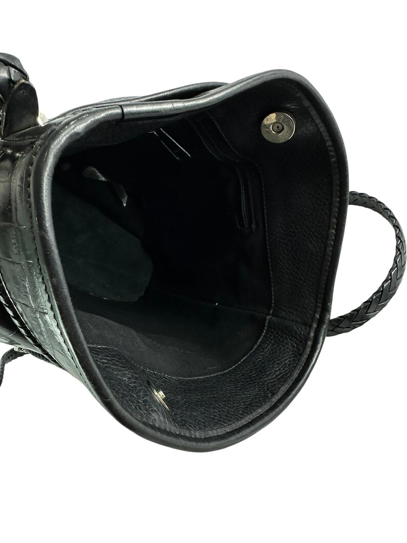 May include: Black leather handbag with a textured crocodile pattern. The interior is black with a small zippered pocket. The bag has a braided handle and a magnetic closure.