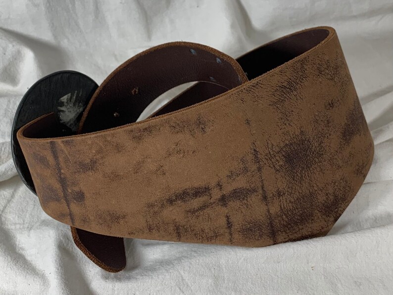 May include: A brown leather belt with a large, round, black buckle. The belt is distressed and has a worn look.