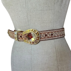 May include: A brown leather belt featuring a geometric pattern of white lines and diamonds. The belt includes a large, oval-shaped gold-tone buckle adorned with small clear crystals and a central faceted, multi-colored reflective stone.