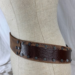 May include: A brown leather belt with metal studs. The belt is worn on a fabric-covered mannequin.