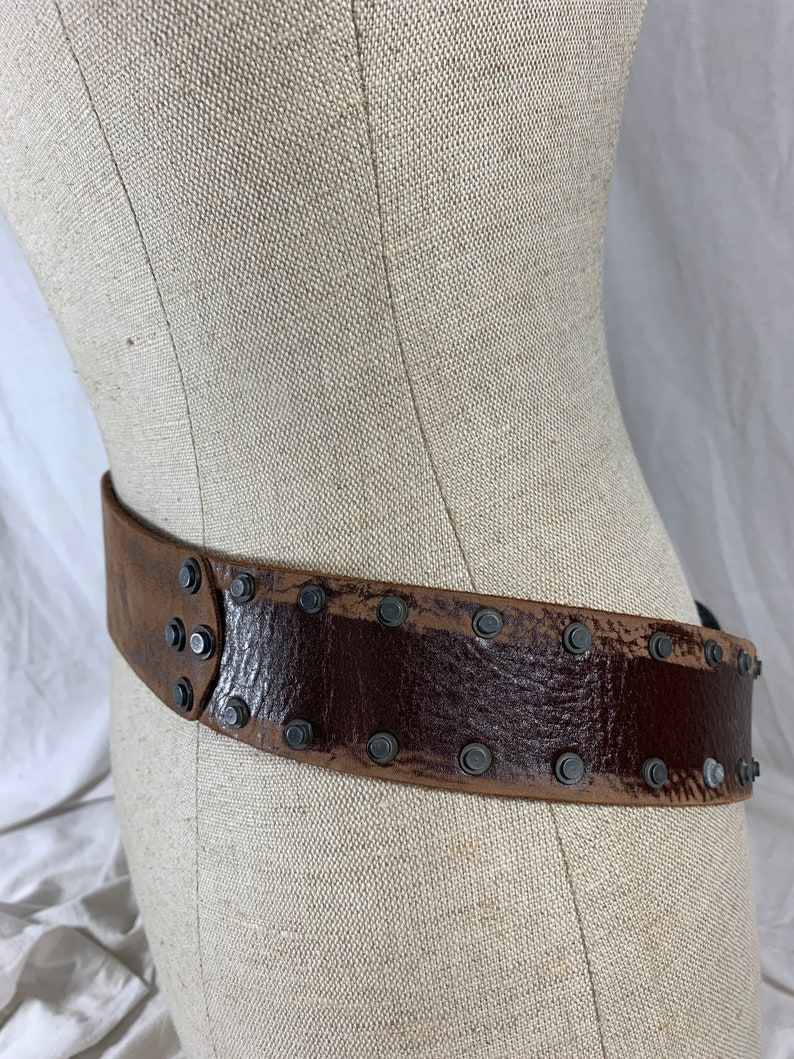 May include: A brown leather belt with metal studs. The belt is worn on a fabric-covered mannequin.