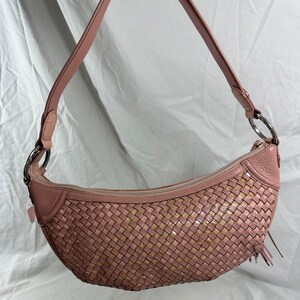 May include: A pink woven shoulder bag with a curved shape. The bag features a woven pattern with gold accents, a zipper closure, and a pink leather strap with silver metal rings. A small tassel hangs from the side.