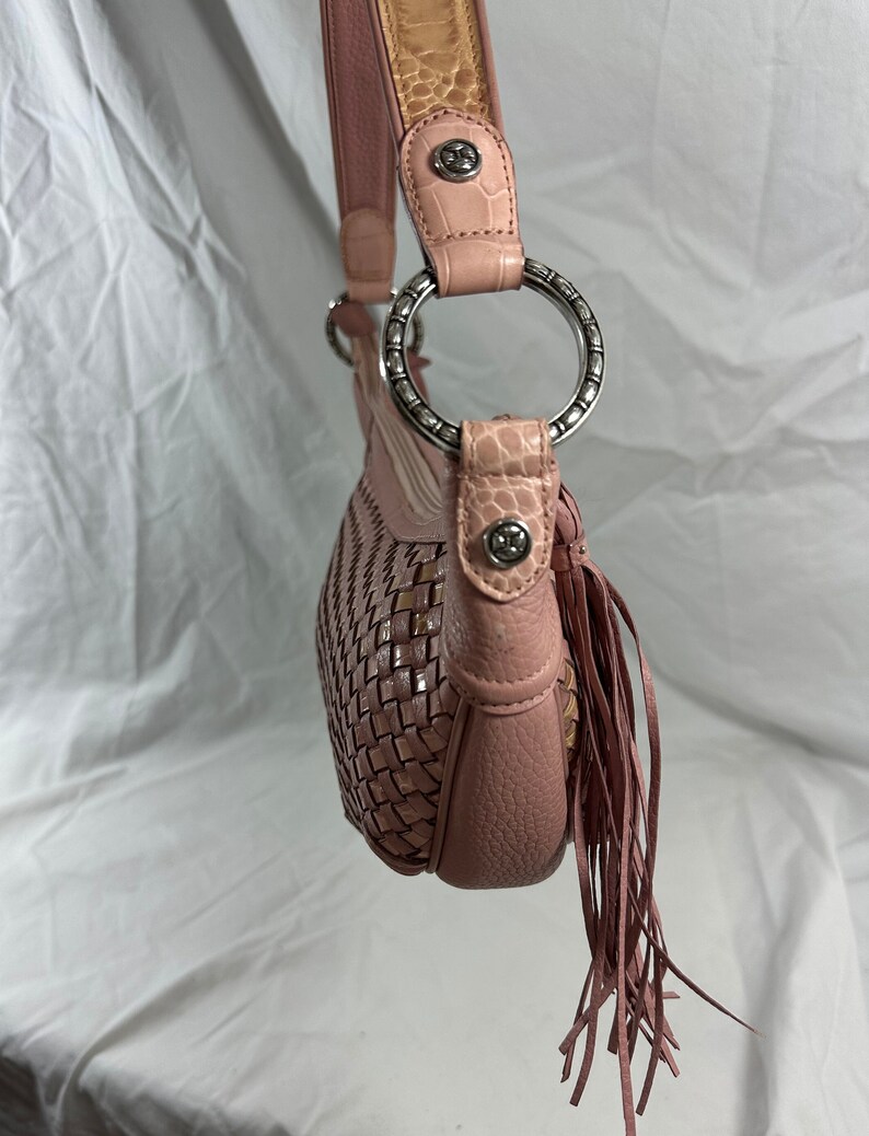 May include: A pink shoulder bag with a woven design on the front. The bag features a leather strap with silver metal rings and decorative fringe. The strap has a light brown crocodile-textured leather accent.