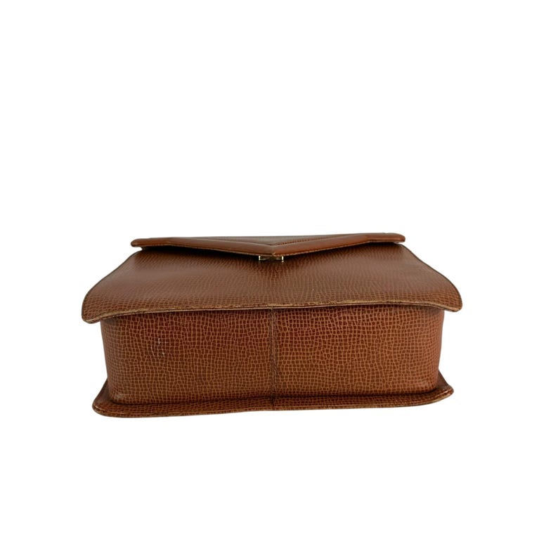 Op de afbeelding: Een bruine leren handtas met een getextureerd oppervlak. De tas heeft een rechthoekige vorm met een klepsluiting en een goudkleurige sluiting. De tas wordt van opzij bekeken.
