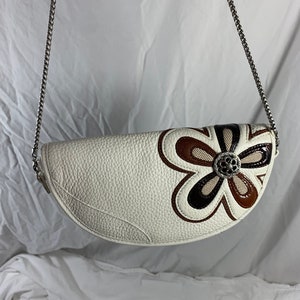 May include: A white leather handbag with a brown and black flower design. The bag has a silver chain strap.