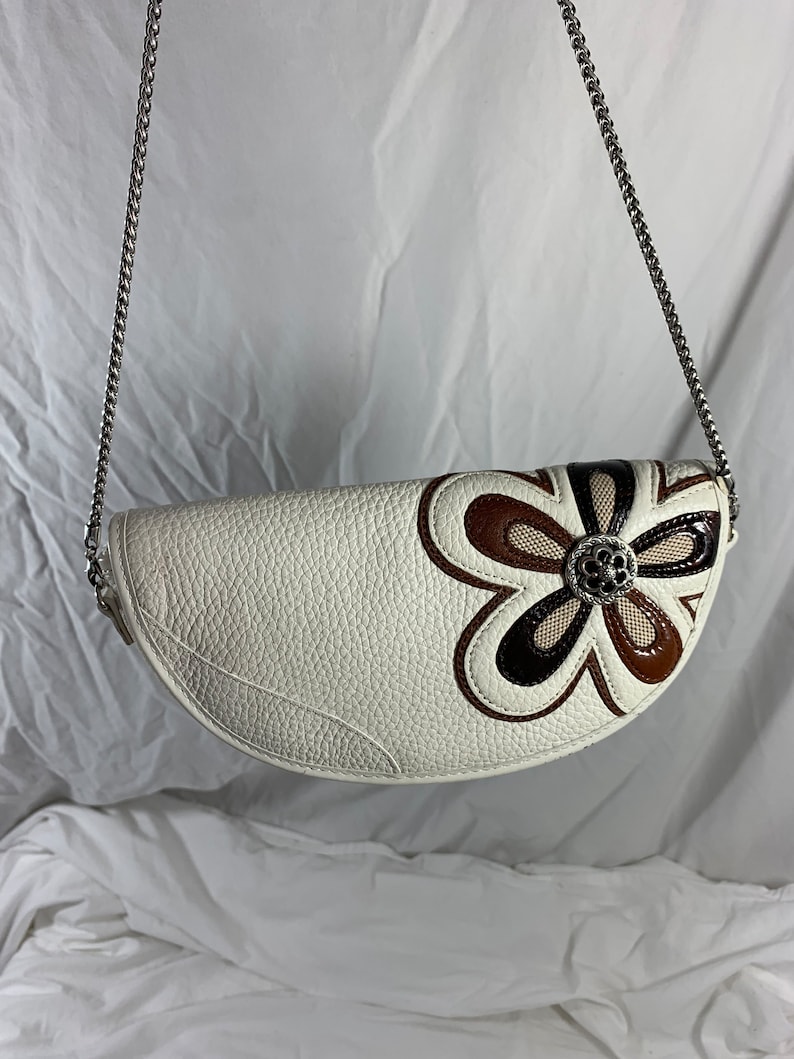 May include: A white leather handbag with a brown and black flower design. The bag has a silver chain strap.