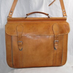 May include: A tan leather briefcase with two buckles and a shoulder strap. The briefcase has a top handle and is made of a distressed leather.
