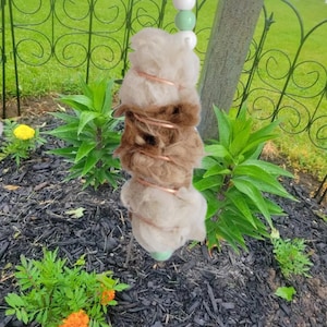 May include: A handmade bird feeder made of natural fibers and copper wire. The feeder is a stacked design with alternating shades of brown and beige, and is suspended from a beaded string. The background includes green plants and a decorative fence.