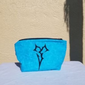 May include: A blue zippered pouch with a black embroidered design. The design is a stylized symbol with sharp points.