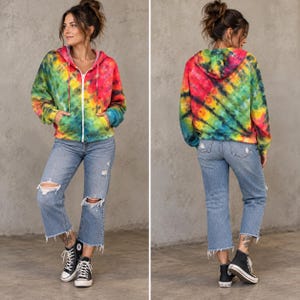 Op de afbeelding: Een hoodie met lange mouwen en rits met een levendig tie-dye patroon in tinten rood, geel, groen en blauw. De hoodie heeft een capuchon met trekkoord, twee voorzakken en een witte ritssluiting, gecombineerd met een lichtblauwe spijkerbroek.