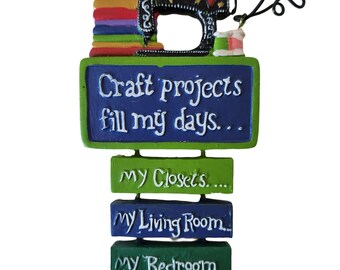 May include: A blue and green sign with the text "Craft projects fill my days... My Closets... My Living Room... My Bedroom..."  A colorful stack of fabric and a black sewing machine with a red heart are above the sign.