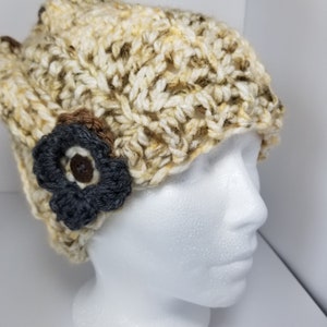 May include: A crocheted beanie hat in beige, brown, and white with a brown and gray flower accent. The hat is on a white mannequin head.