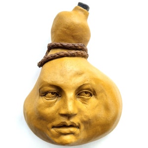 May include: A ceramic gourd sculpture with a human face. The gourd is painted in a yellow ochre color and has a rope tied around the top. The face has a serious expression with closed lips and slightly furrowed brows.