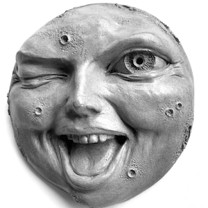May include: A close-up of a gray, sculpted moon face with a winking eye and a wide, toothy grin. The moon face has a textured surface with craters.