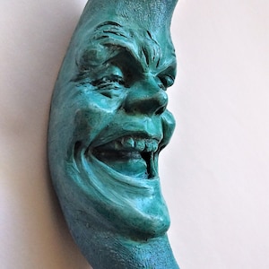 Pode incluir: Uma escultura de lua crescente de cor verde-azulada com um rosto sorridente e dentes afiados.