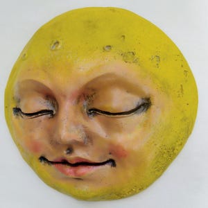 Puede incluir: Una placa de pared circular con forma de luna y rostro humano. La luna está pintada en tonos amarillos y bronceados, con ojos cerrados, una sonrisa suave, mejillas sonrosadas y una superficie texturizada con pequeños cráteres en la parte superior.