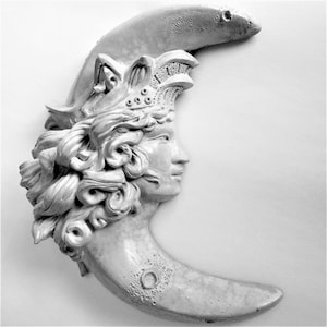 May include: A white ceramic sculpture of a woman's face with flowing hair, set against a crescent moon shape.