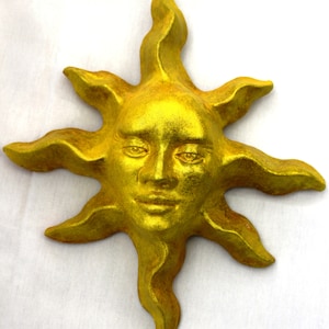 May include: A gold-painted sun sculpture with a human face. The sun has a stylised, wavy design with a face that has closed eyes and a slight smile.