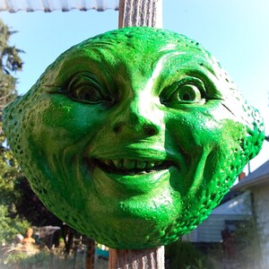May include: A green, ceramic sculpture of a face with a wide, toothy grin. The sculpture is shaped like a lemon and has a textured surface.