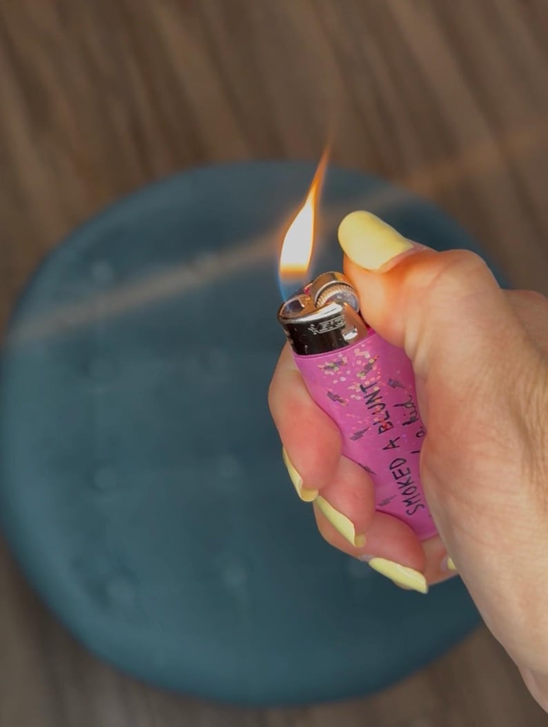 May include: A pink lighter with the text "SMOKED A BLUNT" is held in a hand with yellow nail polish. The lighter has a flame burning. The background is a blurred blue object and wooden surface.
