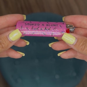 May include: A pink lighter with the text "SMOKED A BLUNT had a kid" in black script. The lighter has a silver metal top and a red ignition button. The background is a blurred teal color.