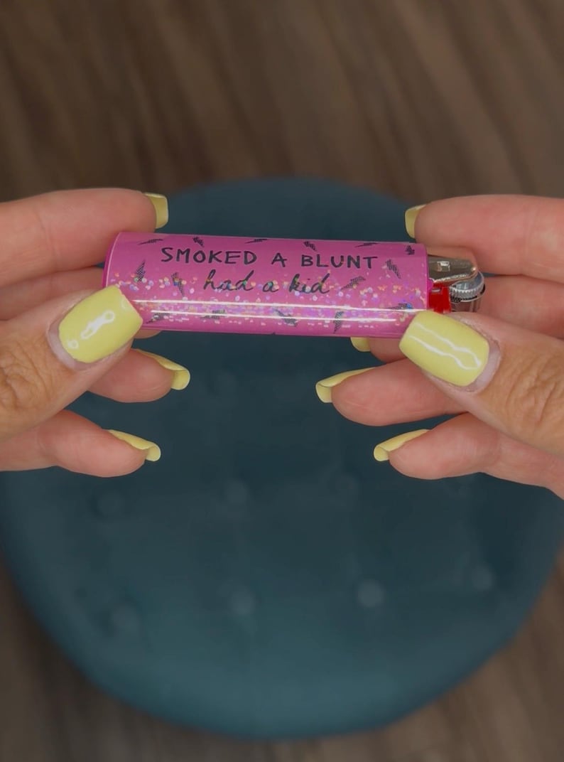 May include: A pink lighter with the text "SMOKED A BLUNT had a kid" in black script. The lighter has a silver metal top and a red ignition button. The background is a blurred teal color.