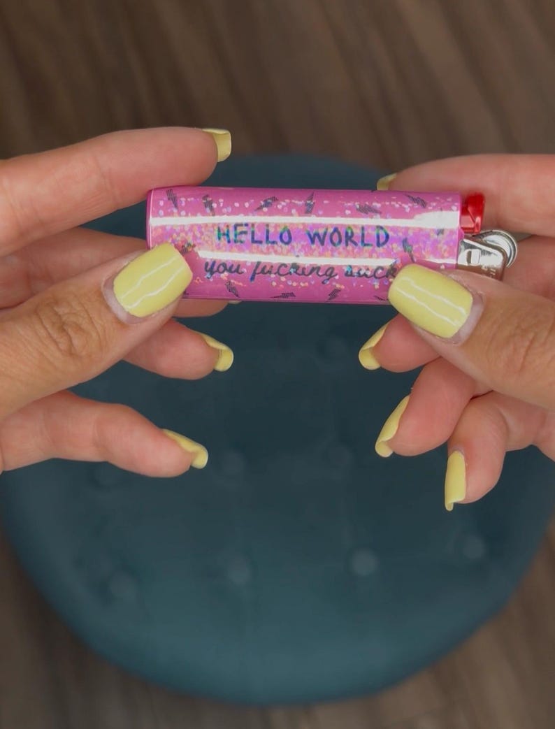 May include: A pink lighter with the words "HELLO WORLD you f*cking suck" printed on it. The lighter has a red ignition button and a silver metal tip. The background is a dark teal color.