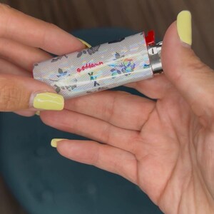 May include: A silver lighter with a floral pattern and the word "goddamn" in pink script. The lighter has a red ignition button and is held between two hands with yellow nail polish.