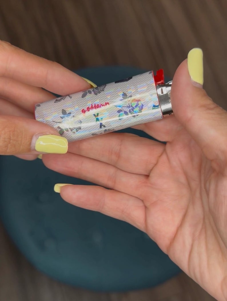 May include: A silver lighter with a floral pattern and the word "goddamn" in pink script. The lighter has a red ignition button and is held between two hands with yellow nail polish.