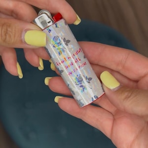 May include: A silver lighter with a floral design and the text "I art my casket, laced with hans & roses." The lighter has a red ignition button and is held by hands with yellow nail polish.