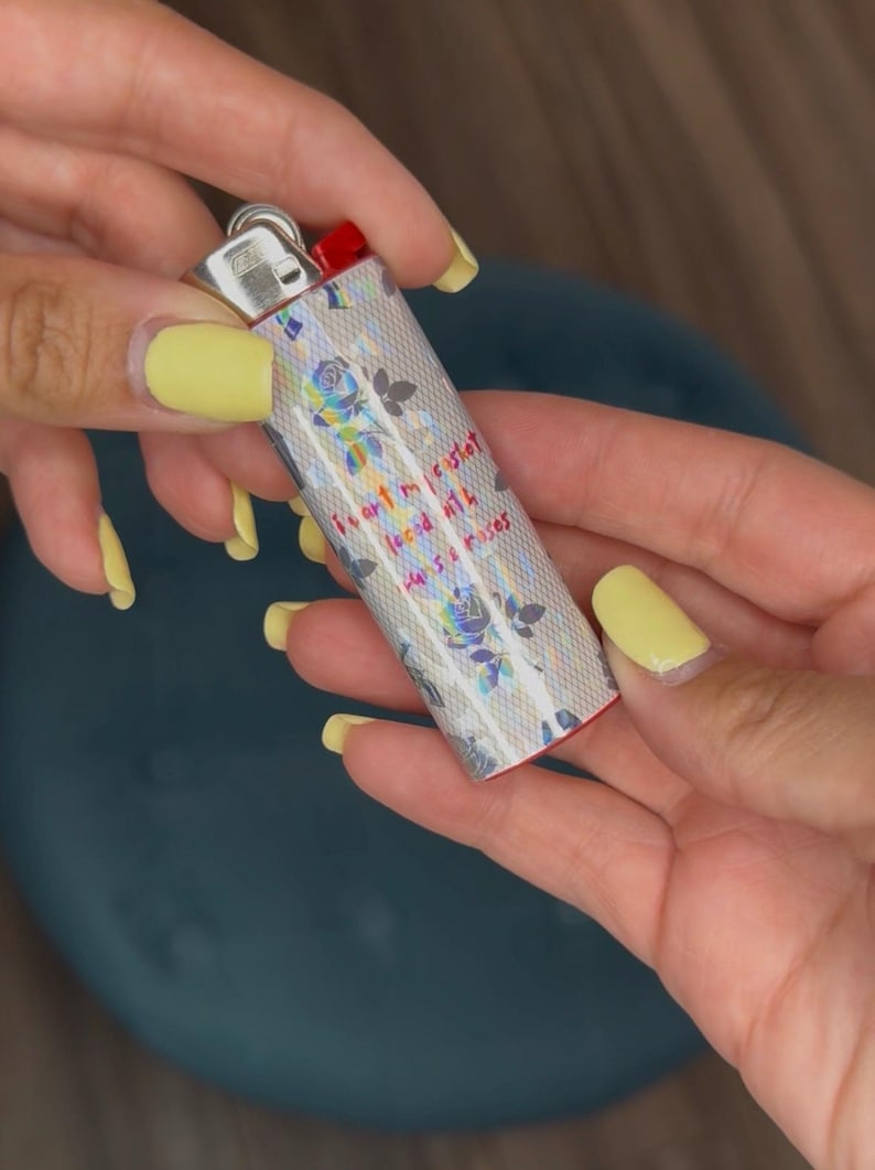 May include: A silver lighter with a floral design and the text "I art my casket, laced with hans & roses." The lighter has a red ignition button and is held by hands with yellow nail polish.