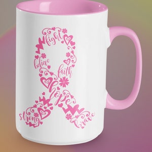 May include: White ceramic coffee mug with a pink rim and handle. The mug features a pink ribbon design with the words "fight", "believe", "faith", "hope", "strong", and "brave" incorporated into the ribbon.