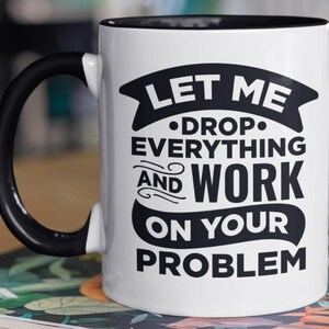 May include: White ceramic mug with a black handle and rim. The mug features the text "LET ME DROP EVERYTHING AND WORK ON YOUR PROBLEM" in black, stylized lettering. The mug is likely used for coffee or tea.