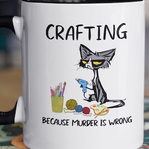 May include: White ceramic mug with a black rim featuring a cartoon cat holding a glue gun. The cat is surrounded by crafting supplies, including yarn, paintbrushes, and a paint palette. The text "CRAFTING BECAUSE MURDER IS WRONG" is printed on the mug.