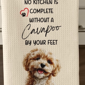 May include: Cream-colored kitchen towel with the text "NO KITCHEN IS COMPLETE WITHOUT A Cavapoo BY YOUR FEET." A smiling Cavapoo dog illustration is at the bottom. The towel has a textured waffle weave.