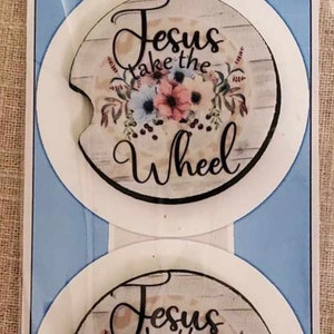 May include: Two car coasters with a floral design and the words "Jesus Take the Wheel." The coasters are white with a floral and wood grain background. The packaging includes the text "Car Coasters" and the website giftsbyapersonaltouch.com.