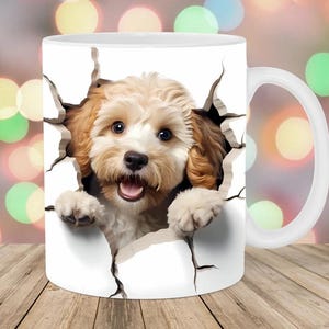 May include: White ceramic mug featuring a playful illustration of a light brown and white dog bursting through a torn surface. The dog has a happy expression with its tongue out. The mug has a white handle and is set against a blurred background.
