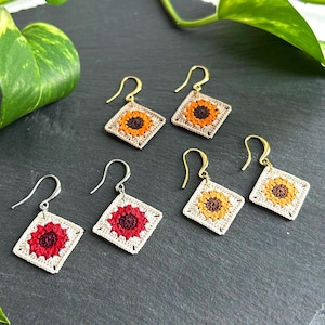 May include: Crochet earrings with a sunflower design. The earrings are square, with a cream border and a yellow, orange, and brown sunflower. The hooks are gold and silver coloured. The earrings are handmade.