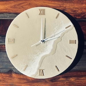 May include: Round wall clock with a beach-themed design. The clock face features a sandy texture with a wave-like pattern. Roman numerals and stick markers are in a light brown color. The clock hands are white.