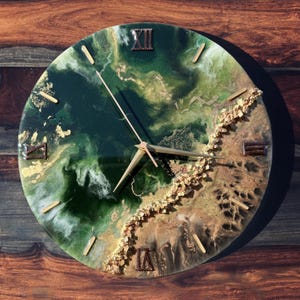 May include: Round wall clock with a unique design. The clock face features a blend of green, gold, and white resin with gold accents. Roman numerals mark the hours. The clock hands are gold. The clock measures approximately 12 inches in diameter.